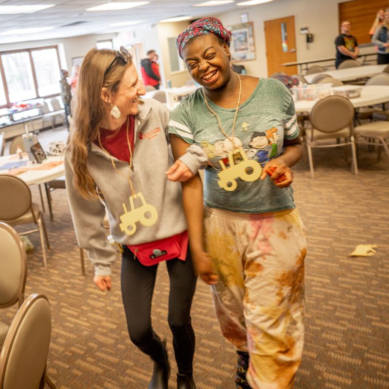 Two women walk arm-in-arm through a classroom, laughing.