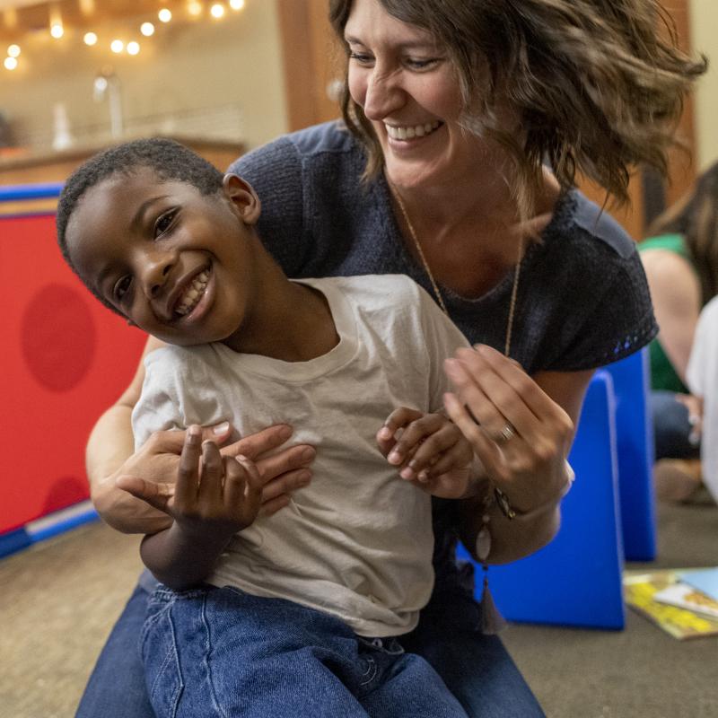 White female therapist joyfully interacts with a dark-skinned boy in a classroom. 