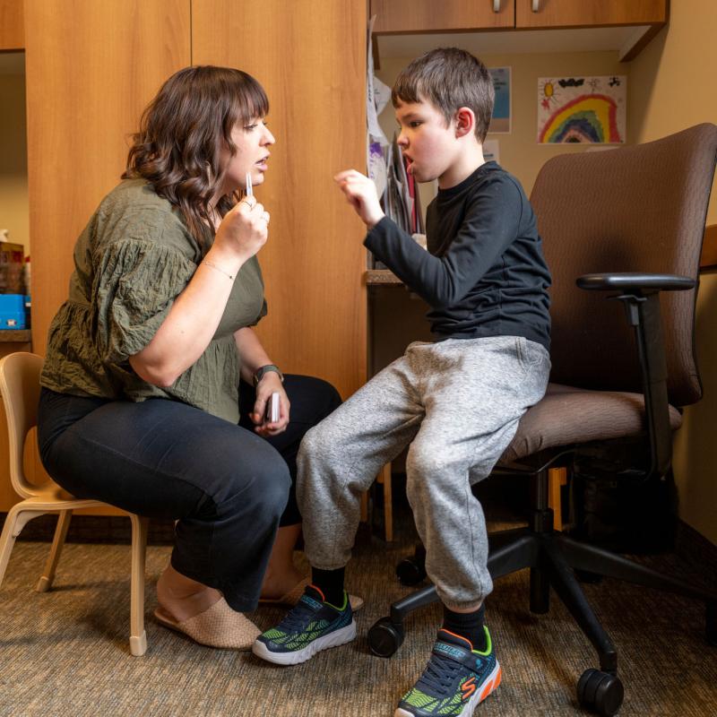 Female therapist and young boy sitting across from each other, mirroring their hand positions and speaking.