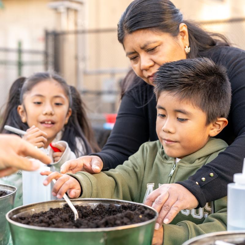 Tan-skinned mother and two children planting herbs at an outdoor event. 